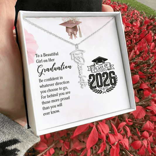 Graduation-themed necklace in a gift box with a message and 'Class of 2026' design, held against a red leafy background.